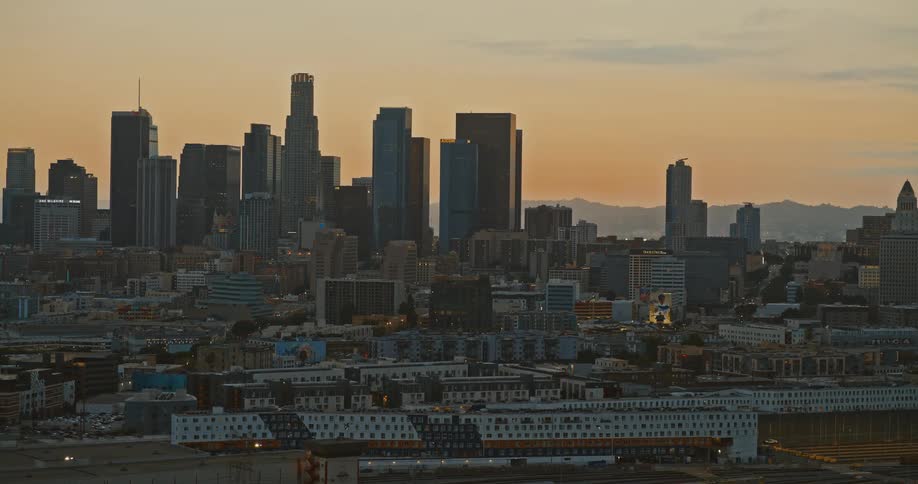 An aerial view slowly zooms in on the Downtown Los Angeles skyline as the sun sets, casting a warm golden glow over the city
