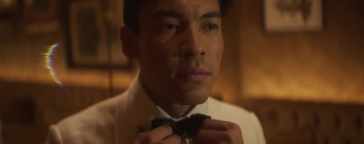 A dapper man in a white tuxedo jacket carefully adjusts his black bow tie in a dimly lit, elegant room