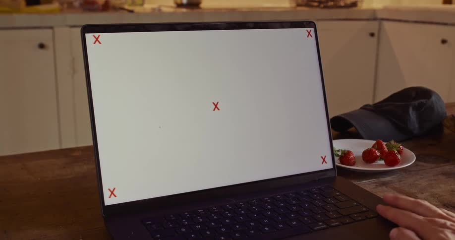 A person rests their hand on the trackpad of a laptop with a trackable blank screen sitting on a wooden table in a kitchen