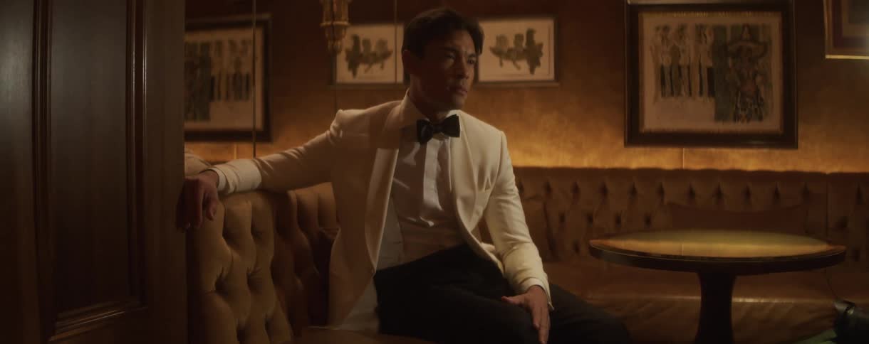 A sophisticated man in a white tuxedo jacket and bow tie sits alone on a plush banquette in a dimly lit, upscale lounge
