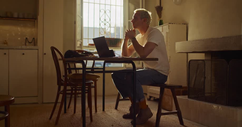A young man with bleached blonde hair works on his laptop at a rustic table, pausing to think in the warm afternoon light