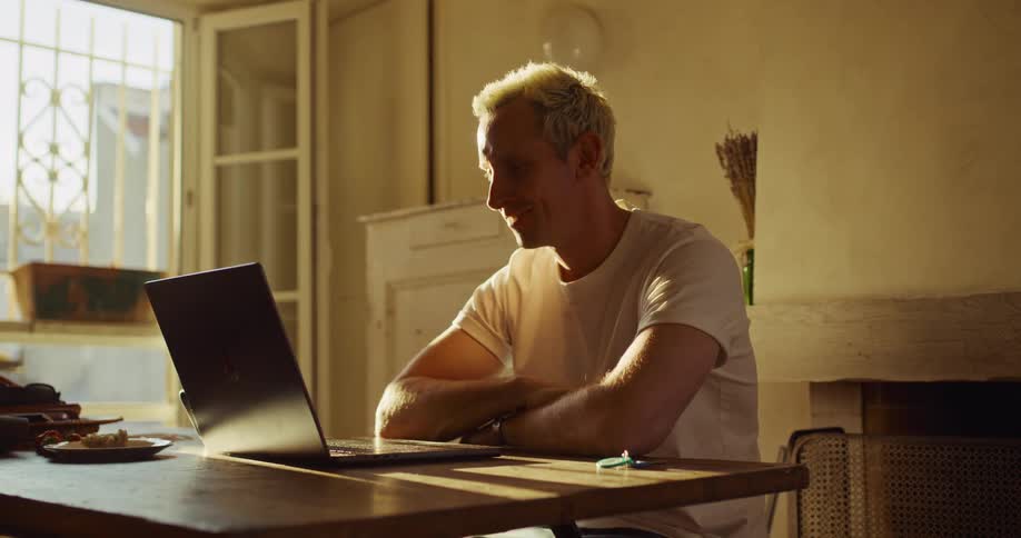 A man with blond hair smiles and waves while on a video call on his laptop in a warm, sunlit room