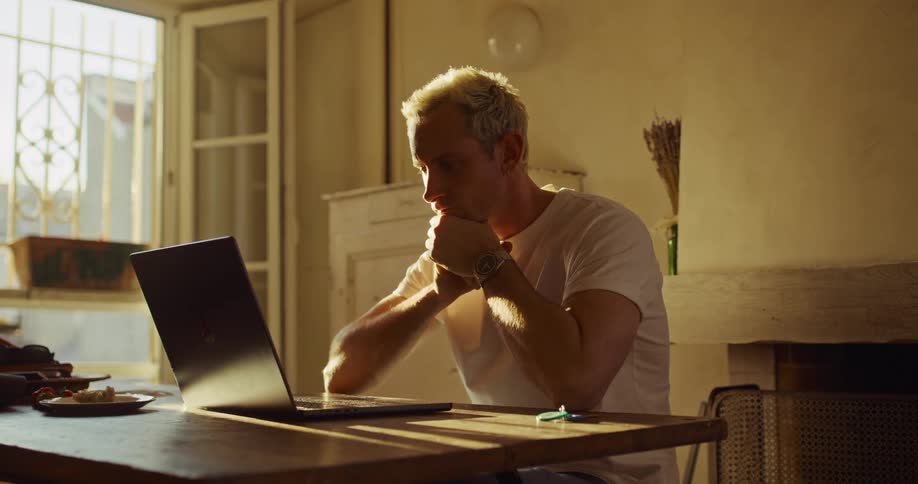 A focused man with bleached blonde hair engages in an expressive video call on his laptop in a warm, sunlit apartment