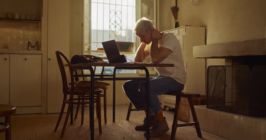 A frustrated young man works from home on his laptop, rubbing his face and neck in a sunlit apartment