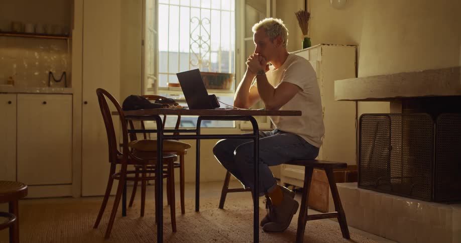 A thoughtful man with platinum blonde hair works on his laptop in a cozy, sun-drenched room, pausing to reflect deeply on his project
