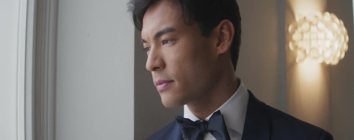 An elegant Asian man in a tuxedo and bow tie thoughtfully looks out a window, preparing for a formal event