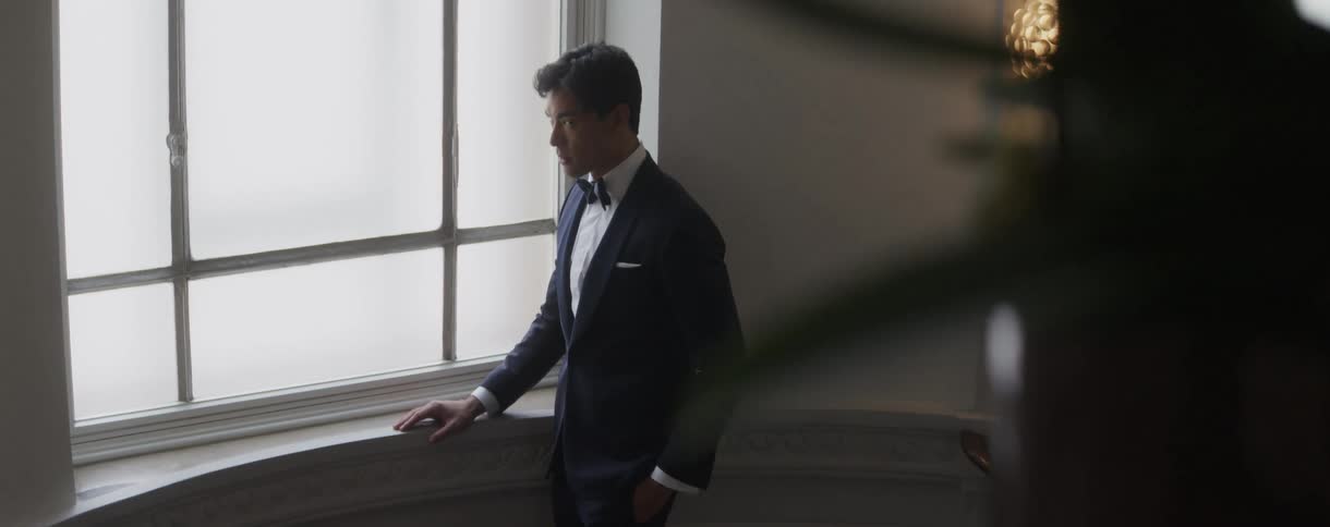 A handsome man in a formal tuxedo stands pensively, looking out a bright window while waiting for an event to begin