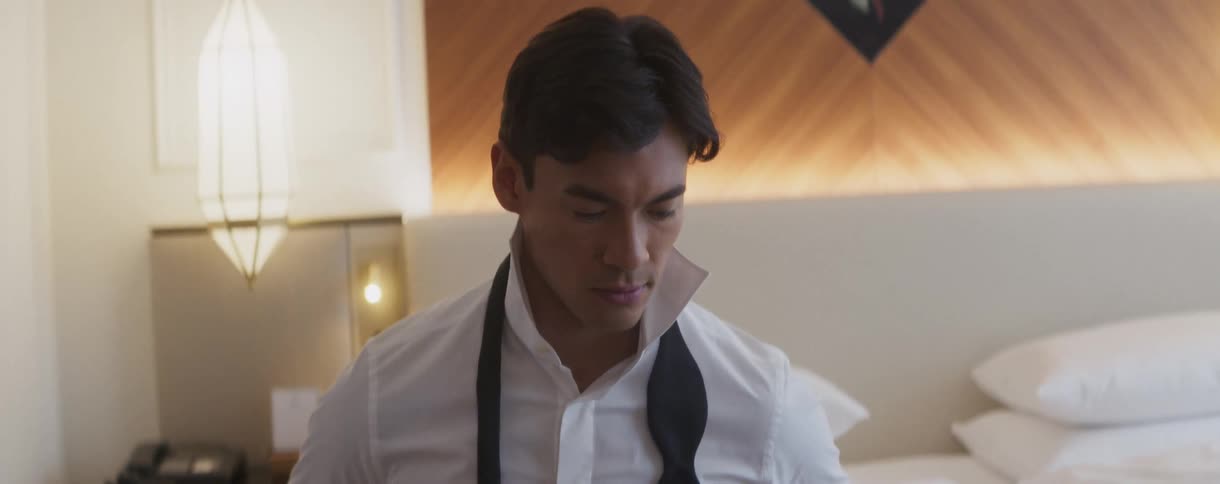 A handsome, well-dressed man sits contemplatively on a bed in a hotel room while preparing for a formal event