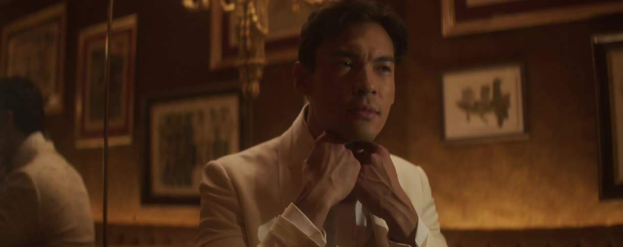 A dapper Asian man in a white tuxedo adjusts his bow tie while looking in the mirror of a luxurious, dimly lit room