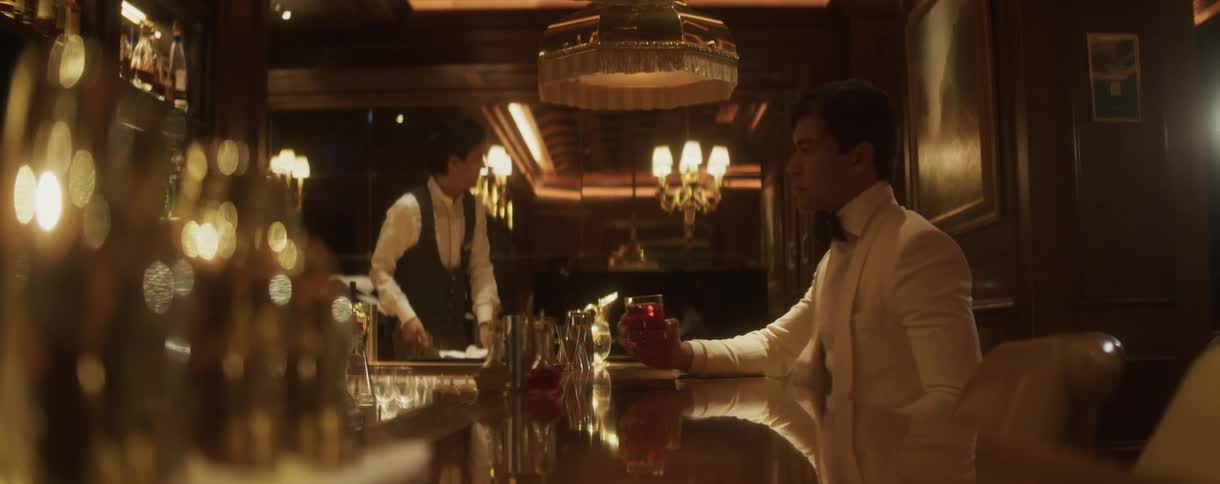 A dapper gentleman in a white tuxedo sits at a sophisticated, dimly lit bar and slowly enjoys a red cocktail