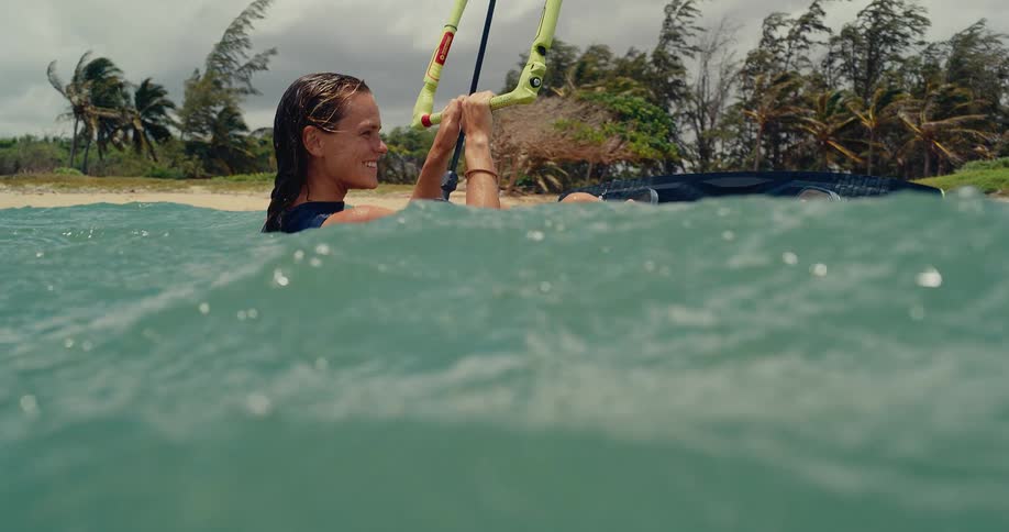 A female kitesurfer sits smiling in the tropical ocean waves, holding her control bar and preparing to ride in slow motion