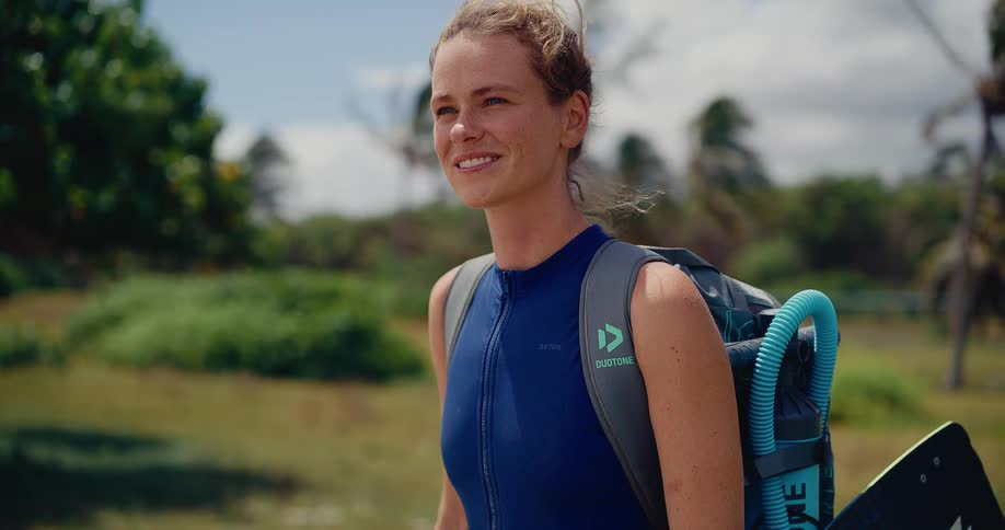 A happy female athlete in a wetsuit, carrying her kitesurfing gear, smiles while looking out at the windy tropical beach