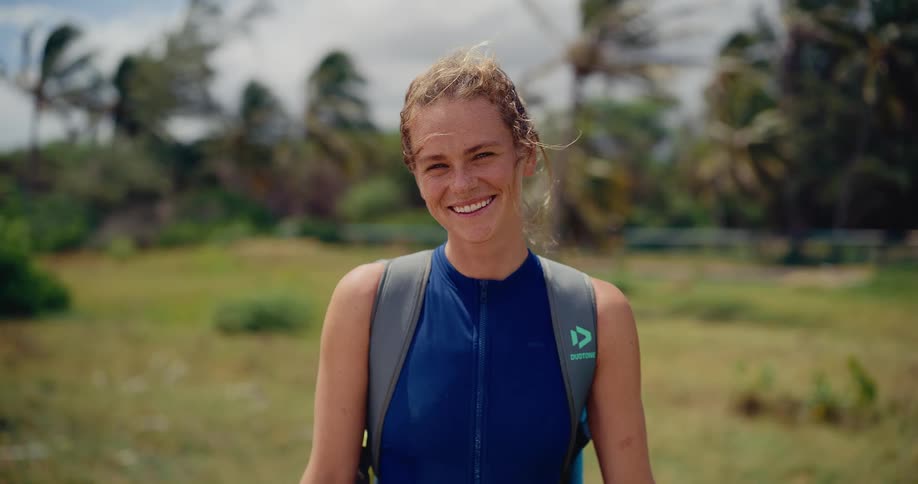 A close-up portrait of a happy athletic woman in a wetsuit smiling at the camera on a windy day near the beach