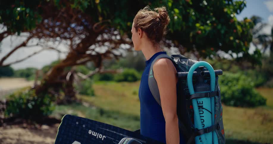 Confident female kitesurfer stands with her board and pack, looking towards the water on a beautiful tropical beach