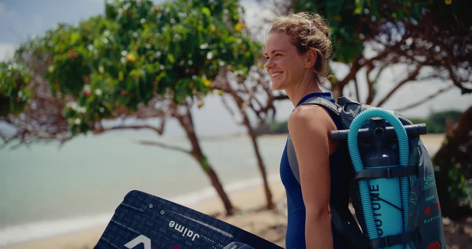 A young female kitesurfer smiles while looking out at the ocean, holding her board on a beautiful tropical beach