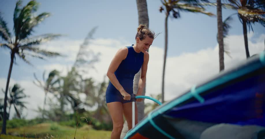 A smiling woman in a wetsuit inflates her kitesurfing kite with a pump on a windy tropical beach with palm trees
