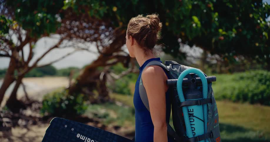 A happy female kitesurfer in a wetsuit smiles at the camera before turning to look out at the beautiful tropical beach, ready for her session