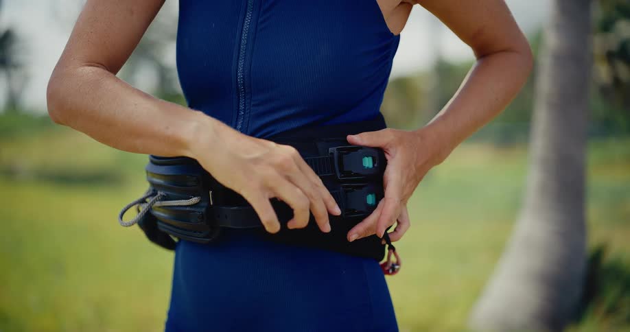 A female athlete in a blue wetsuit meticulously adjusts and tightens her black kitesurfing waist harness in preparation for her session