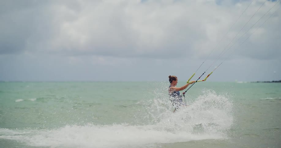 A focused female kitesurfer glides across the ocean surface, creating a trail of sea spray in slow motion