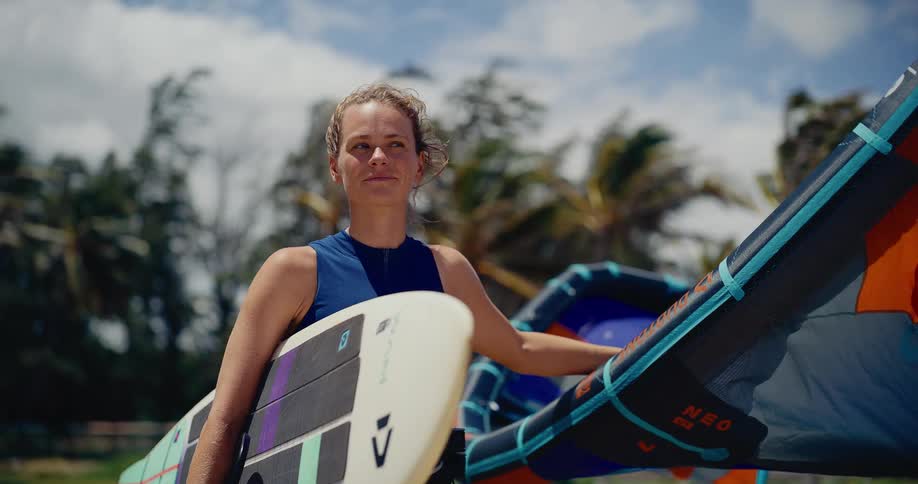 Portrait of a smiling young woman holding her kitesurfing board and kite on a windy tropical beach