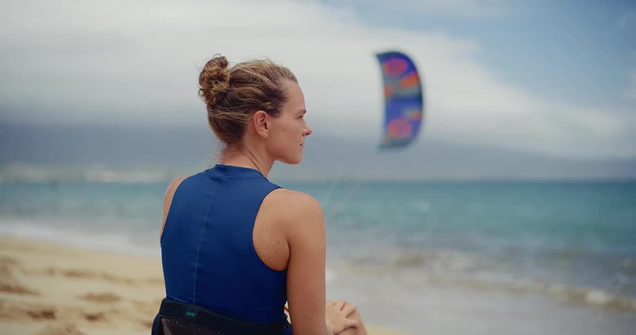 A female kitesurfer sits on the beach in her gear, looking out at her kite over the ocean before her session