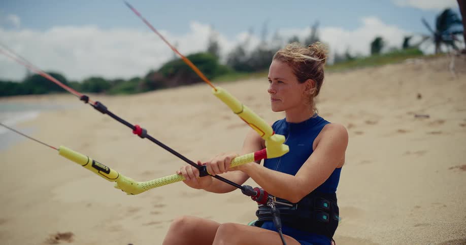 A happy female kitesurfer sits on a sandy beach with her equipment, smiling and getting ready to go out on the ocean