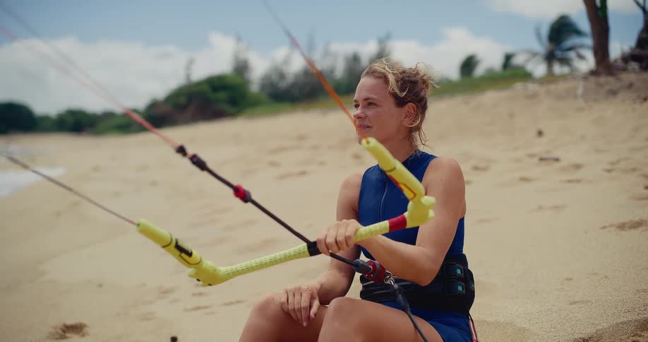 A happy female kitesurfer sits on a sandy beach holding her kite's control bar and smiling before heading into the ocean