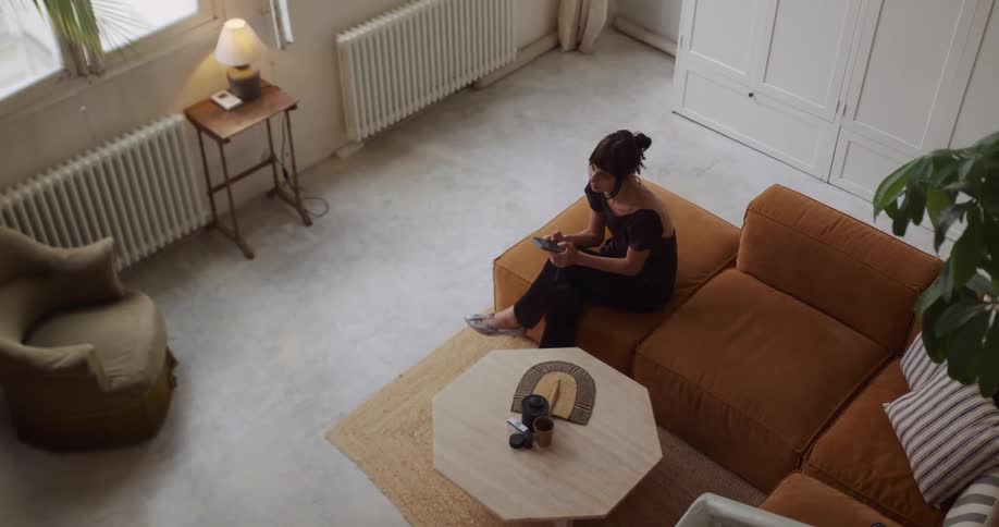 A high-angle view of a woman sitting on a couch, looking at her phone, then getting up and walking away in her stylish apartment