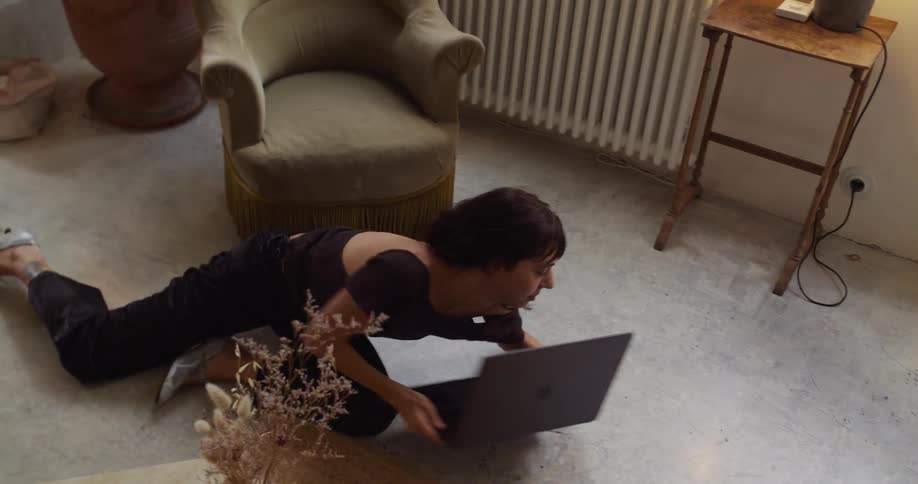 A high-angle shot of a woman comfortably working on her laptop on the floor, changing from lying on her back to her front