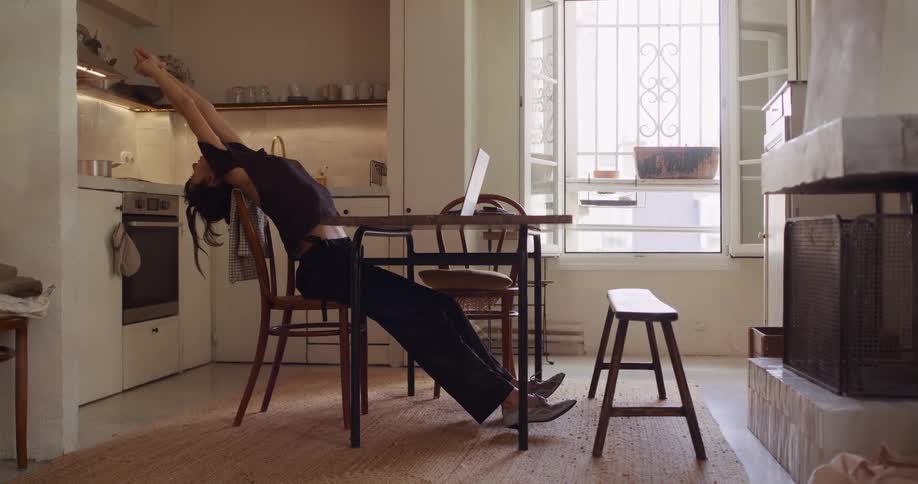 A young woman in a cozy apartment stretches back in her chair before resuming work on her laptop