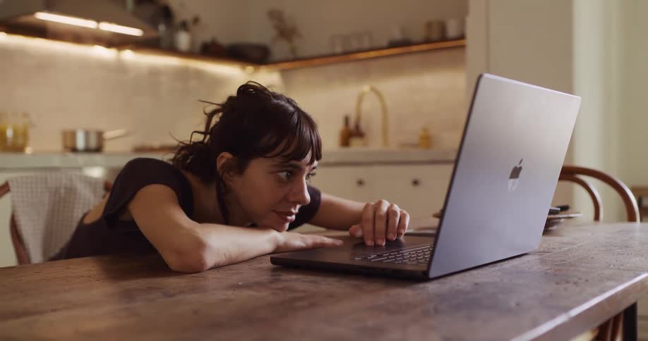 A woman transitions from relaxed browsing to focused work on her laptop at a wooden table in her modern kitchen