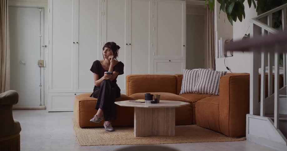 A contemplative woman pauses from scrolling on her phone and looks away thoughtfully while sitting on a modern couch in her living room