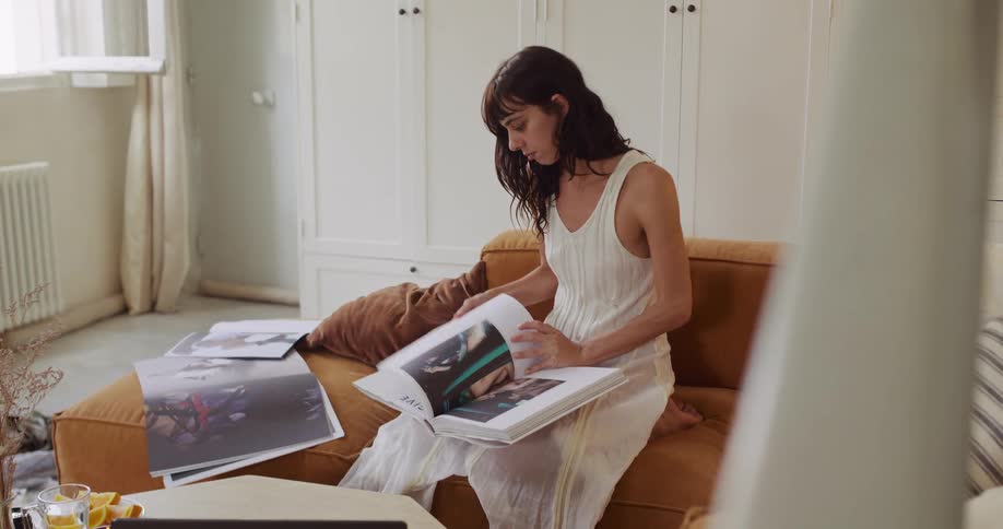 A focused young woman in a white dress sits on her sofa and looks through a large art book in her sunlit, stylish apartment
