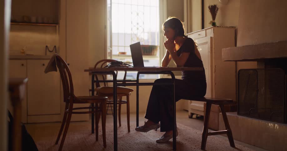 A woman prepares to work from home, tying up her hair and stretching before focusing on her laptop in a sunlit room