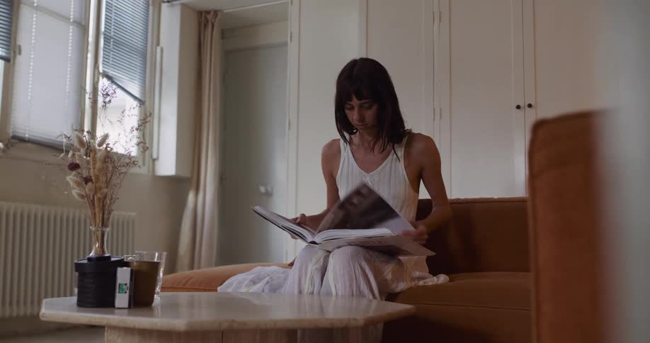 A young woman in a white dress sits on a tan sofa and peacefully flips through a coffee table book in her modern, sunlit apartment