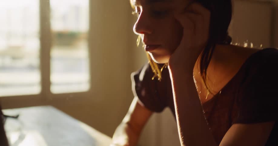 A pensive young woman types on her laptop and then thoughtfully pauses while working in a dusty, sun-drenched room