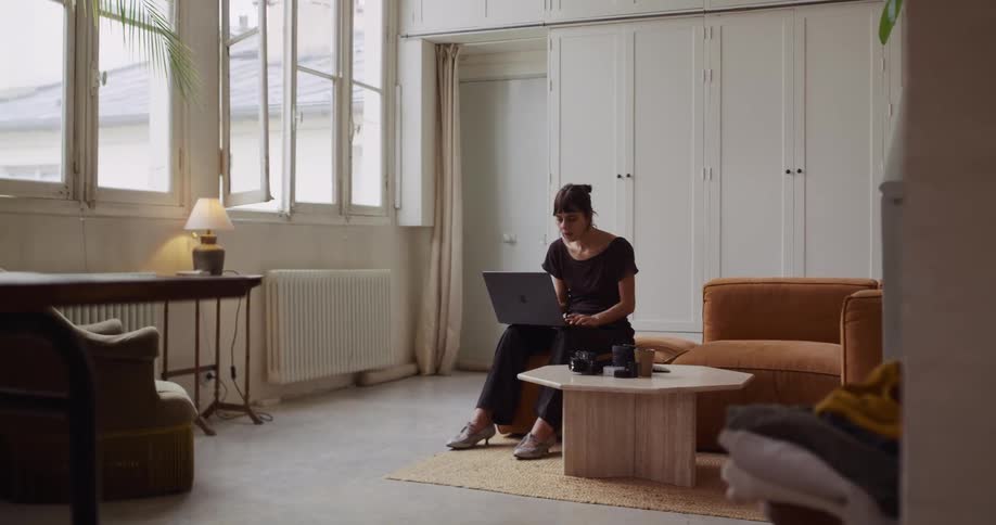 A woman sits on a couch with her laptop, appearing tired or contemplative before beginning to work in her spacious, sunlit living room