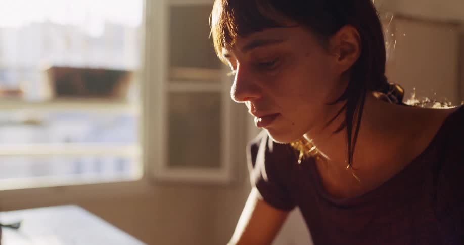 A thoughtful woman intently uses her laptop, bathed in the warm light from a nearby window