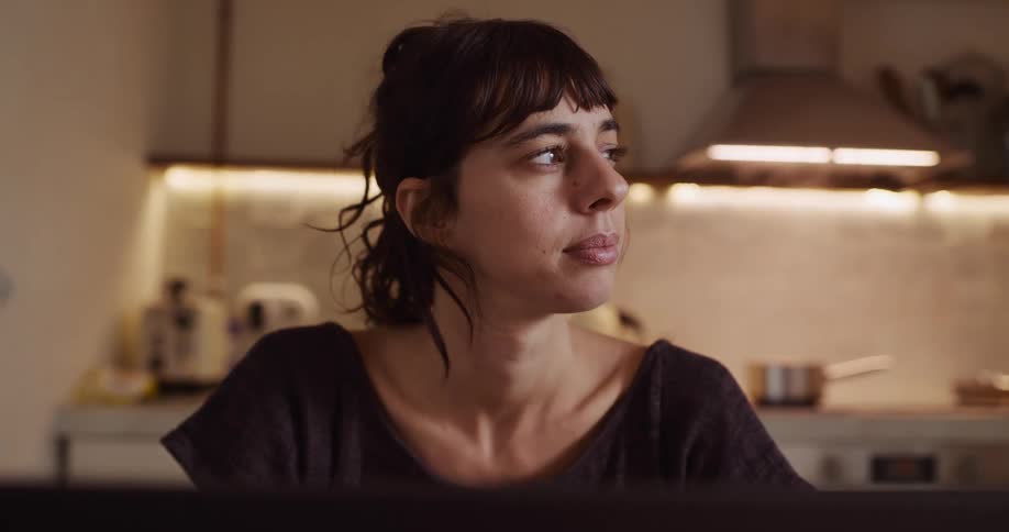 A thoughtful woman working on her laptop at home pauses to look away before returning her attention to the screen