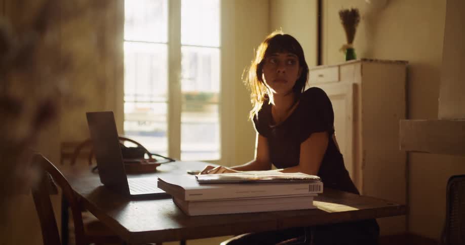 A tired young woman takes a pensive break from her studies, sitting at a sunlit wooden desk with her laptop and books