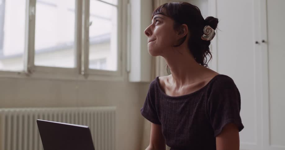 A creative young woman works from home on her laptop, pausing to contemplate before a smile of satisfaction appears