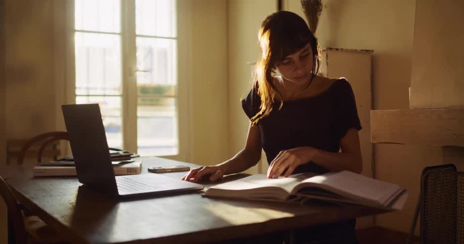 A young woman sits at a table in a warm, sunlit room, working on her laptop and flipping through a book