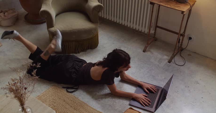 A high-angle view of a relaxed woman lying on the floor of her stylish apartment, working on her laptop and kicking her feet