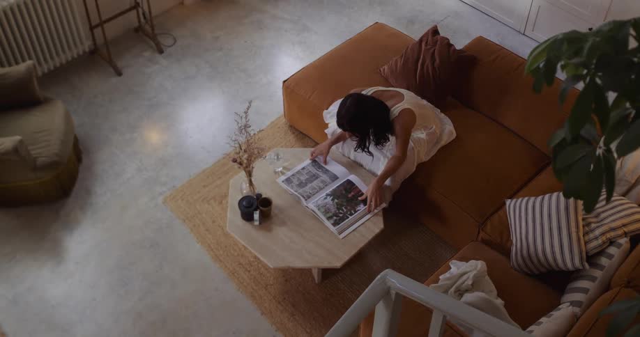 A woman in a flowing white dress walks barefoot through her stylish, earth-toned living room and settles on the couch to look through a large book