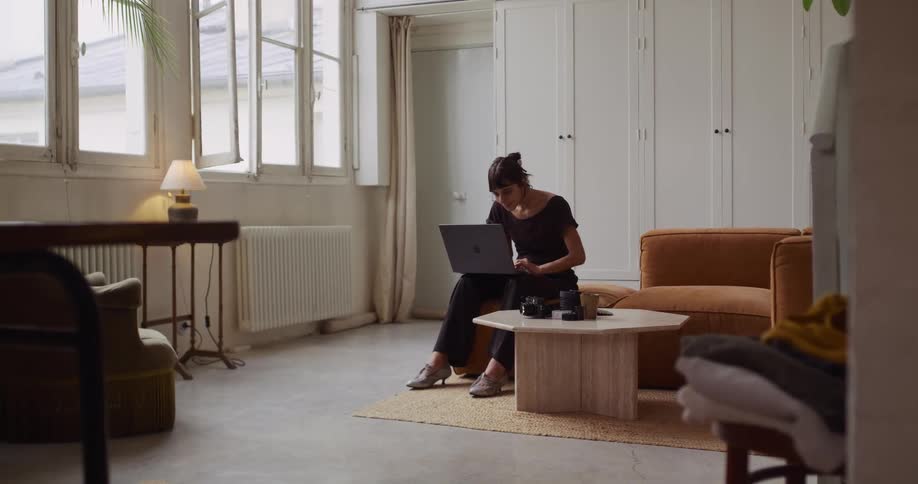 A thoughtful female photographer sits in her stylish, naturally lit apartment and works on her laptop, surrounded by her camera equipment