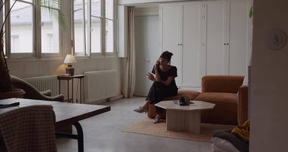 A woman walks through her bright, modern apartment while having a serious conversation on her smartphone, eventually sitting down