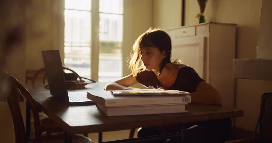 A tired young woman resting her head on a stack of books at a desk wakes up and begins working on her laptop in a sunlit room
