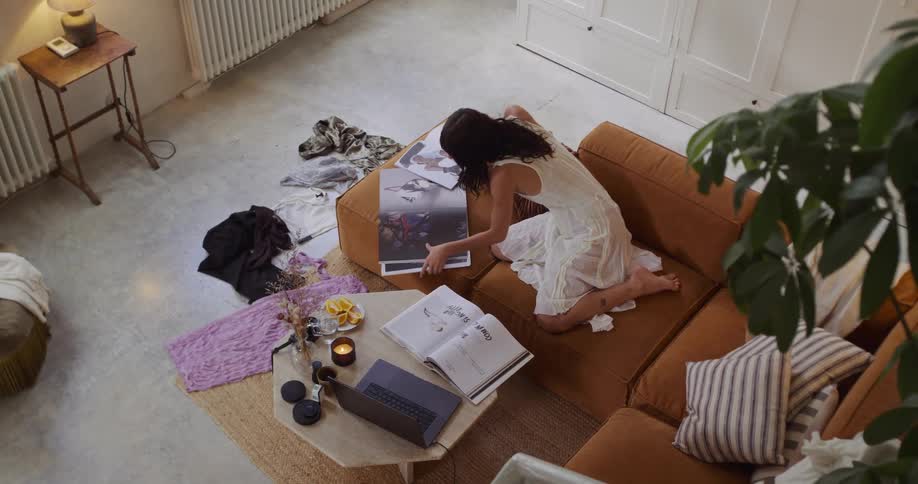 A barefoot woman in a white dress kneels on her couch, drawing inspiration from art prints before working on her laptop in a cozy living room
