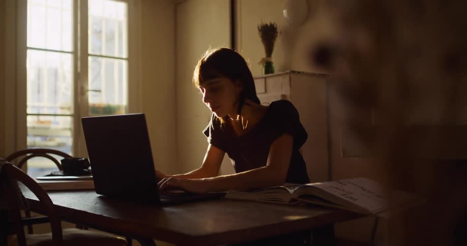 A young woman bathed in the golden hour light works on her laptop at a desk in her cozy apartment