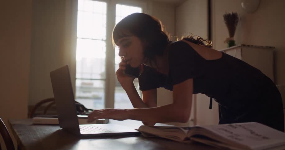 A young woman multitasks, talking on the phone while walking over to use her laptop in a beautifully sunlit apartment